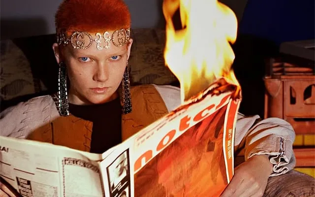 マーランド・バッカスは新聞紙に火を付けている。 彼女は赤い髪をうねらせ、額にヘッドバンドを巻いています。